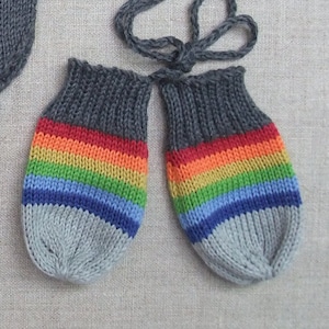 May include: A pair of grey knitted baby mittens with a rainbow stripe design. The mittens are tied together with a grey string.
