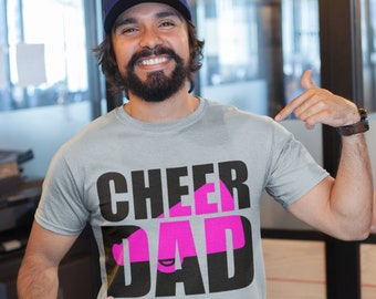 May include: A light grey t-shirt with the words "CHEER DAD" in black and pink. The shirt is being pointed at by a person wearing a blue baseball cap and blue jeans.