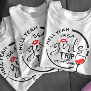 Könnte beinhalten: Drei weiße T-Shirts mit einem schwarz-roten Grafikdesign. Das Design enthält die Worte "HELL YEAH ISSA GIRLS TRIP" in kreisförmiger Anordnung, einen roten Lippenstiftkuss und ein Weinglas. Der Text "CUSTOM DESTINATION HERE" ist ebenfalls enthalten.
