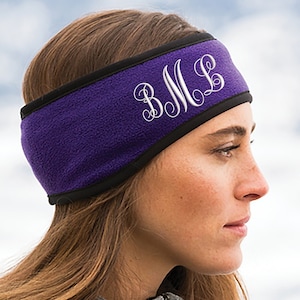 May include: A purple fleece headband with a black trim and white embroidered initials. The headband is worn by a person wearing a gray jacket. The background is blurred, suggesting an outdoor setting.