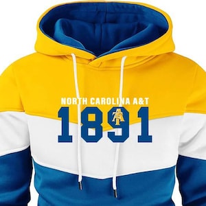 Puede incluir: Una sudadera con capucha en amarillo, blanco y azul con el texto "NORTH CAROLINA A&T" y el n&uacute;mero "1891" en azul marino. La sudadera tiene un cord&oacute;n y un bolsillo tipo canguro.