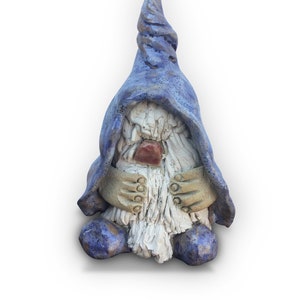 May include: A ceramic garden gnome figurine with a blue pointed hat and matching robe. The gnome has a long white beard, tan hands, and a red nose. The figurine is designed to sit upright.