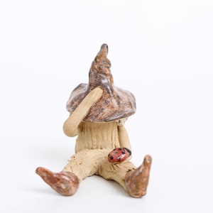 May include: A whimsical ceramic mushroom figurine with a pointed hat and seated pose. The mushroom has a brown and tan speckled hat, a tan body, and brown feet. A small red ladybug with black spots rests on its leg. The figurine is on a white background.