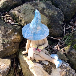 May include: A ceramic fairy figurine with a light blue mushroom cap hat and matching shoes. The fairy is seated on a rock, holding a small bouquet of colourful flowers and a butterfly. The figurine has a whimsical design.