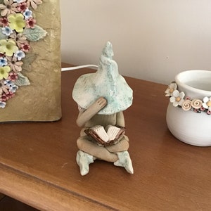 May include: A ceramic gnome figurine, seated and reading a book, with a light blue and white mushroom hat. The gnome's face is hidden. The figurine is on a wooden surface. Other ceramic items are in the background.