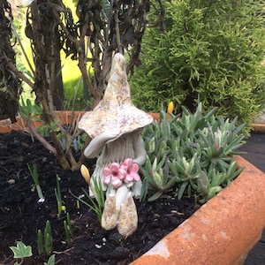 May include: A ceramic gnome figurine with a mushroom hat and pink flowers, set in a terracotta pot with green plants and dark soil. The gnome is light beige with brown speckles. The pot is filled with plants.