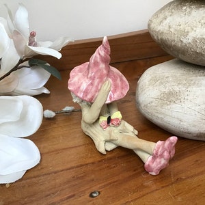 May include: A ceramic fairy figurine, seated with a pink hat and shoes. The fairy has a butterfly detail and rests on a wooden base. White magnolia flowers and decorative stones are in the background.