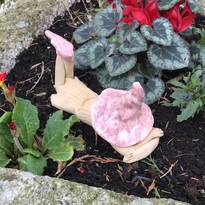 May include: A decorative garden gnome with a pink hat and light brown body, positioned amongst green plants and red flowers. The gnome is in a reclining pose, bringing a playful element to the garden.