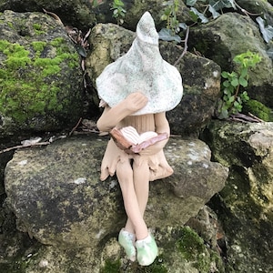 May include: A ceramic fairy figurine with a pointed hat and a book, seated on a moss-covered stone wall. The fairy wears a light green hat and shoes, with a tan dress. The book has pink pages.