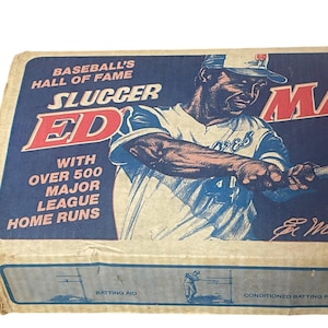 May include: A vintage cardboard box for a baseball game called "Slugger Ed" featuring a baseball player in a blue and white uniform swinging a bat. The box has the text "Baseball's Hall of Fame" and "With Over 500 Major League Home Runs".