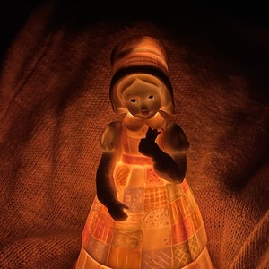 May include: A ceramic figurine of a girl wearing a patchwork dress and a hat. The figurine is illuminated from below, casting a warm glow on the girl's face and dress.