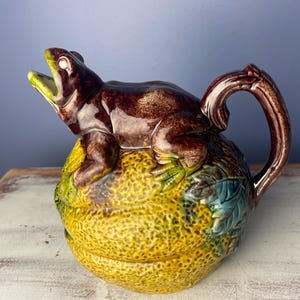 May include: A ceramic pitcher with a frog-shaped lid. The pitcher is brown and yellow with green accents. The frog is brown with a yellow mouth. The handle is brown and curved. The pitcher is likely vintage.