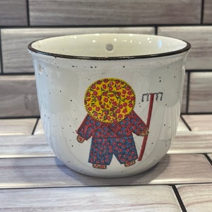 May include: White ceramic hanging planter with a brown rim and a colorful illustration of a person wearing a yellow hat and red and blue clothing holding a rake. The planter has drainage holes.