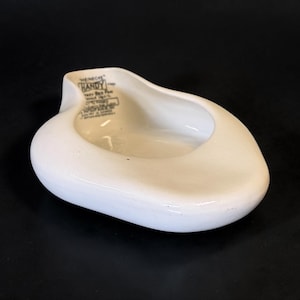 May include: A white ceramic bed pan with a smooth, contoured shape. The interior features black printed text that reads Heinecke Handy Bed Pan, along with additional patent and manufacturing information. The object has a wide, open basin.