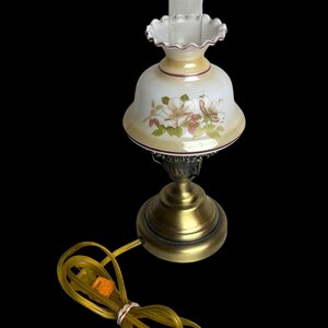 May include: A vintage table lamp with a floral design on the glass shade. The lamp has a brass base and a clear glass chimney. The shade is white with pink and green floral accents. The lamp's cord is yellow.