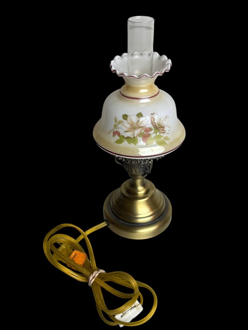 May include: A vintage table lamp with a floral design on the glass shade. The lamp has a brass base and a clear glass chimney. The shade is white with pink and green floral accents. The lamp's cord is yellow.