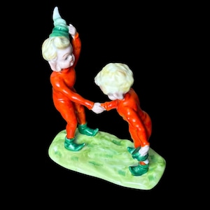 May include: A porcelain figurine featuring two figures with blonde hair wearing orange jumpsuits and green pointed shoes. The figures are holding hands while standing on a textured green base. One figure wears a green pointed hat, while the other is bareheaded.