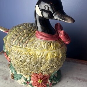 May include: A ceramic goose-shaped serving dish with a red bow around its neck. The goose is decorated with green leaves, pine cones, and a red poinsettia.