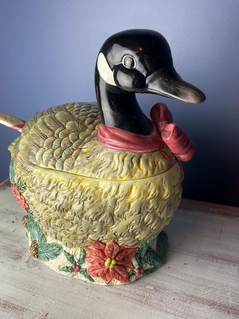 May include: A ceramic goose-shaped serving dish with a red bow around its neck. The goose is decorated with green leaves, pine cones, and a red poinsettia.