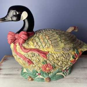 May include: A ceramic goose-shaped serving dish with a red bow and green and brown foliage details. The dish is decorated with a festive holiday theme.