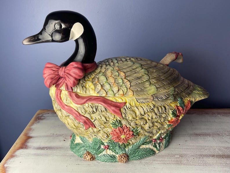 May include: A ceramic goose-shaped serving dish with a red bow and green and brown foliage details. The dish is decorated with a festive holiday theme.