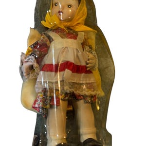 May include: A vintage porcelain doll in a clear plastic package. The doll wears a yellow headscarf, a floral print dress, and a white apron with red stripes. It has black shoes and white socks. The doll is holding a small bag.