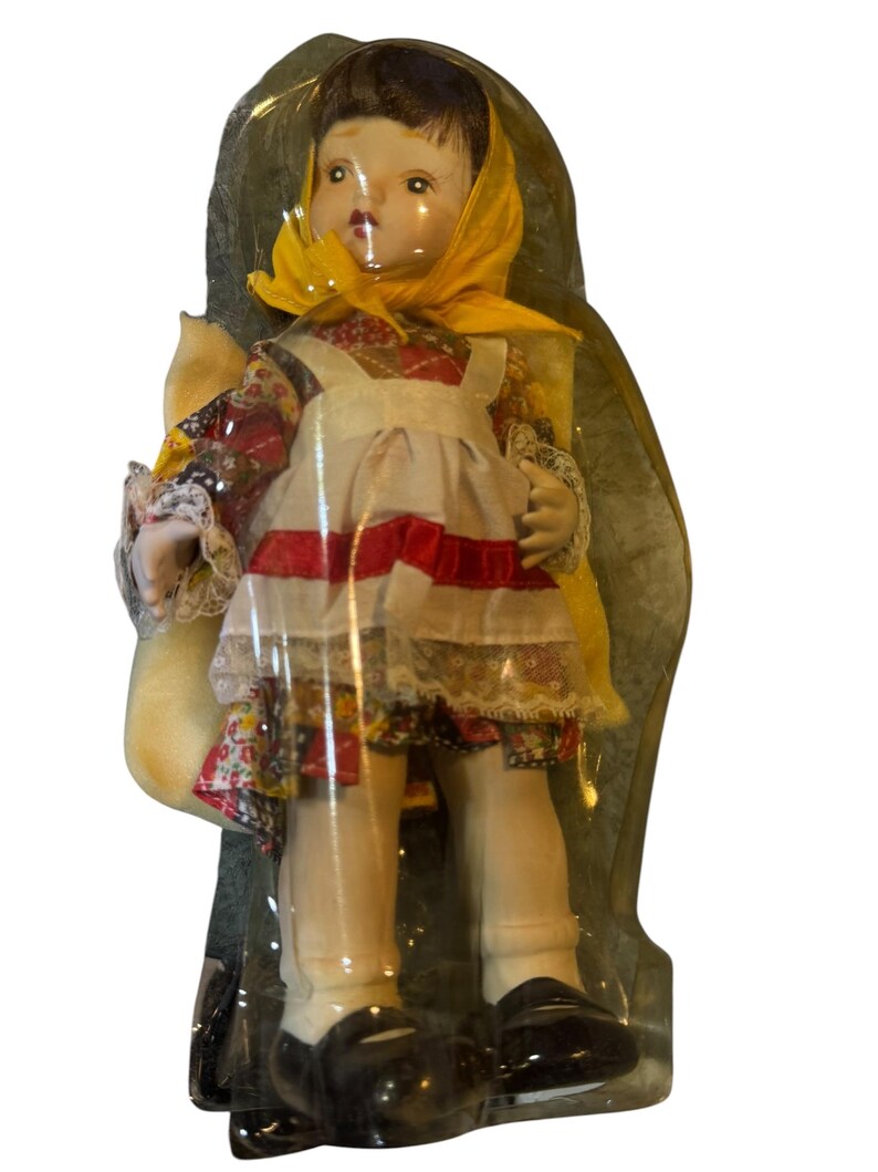 May include: A vintage porcelain doll in a clear plastic package. The doll wears a yellow headscarf, a floral print dress, and a white apron with red stripes. It has black shoes and white socks. The doll is holding a small bag.