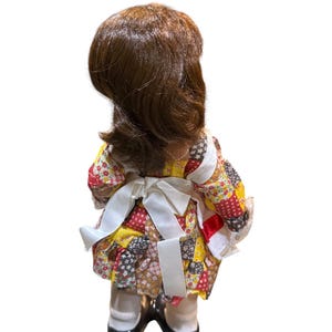 May include: A vintage doll with brown hair and a patchwork dress in red, yellow, and brown. The doll wears a white apron tied in a bow at the back and black shoes. The doll is standing with its back to the viewer.