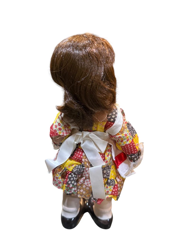May include: A vintage doll with brown hair and a patchwork dress in red, yellow, and brown. The doll wears a white apron tied in a bow at the back and black shoes. The doll is standing with its back to the viewer.