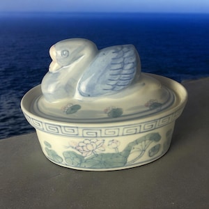May include: A ceramic trinket box with a swan-shaped lid. The swan is white and blue, with detailed wing patterns. The box is white with blue and green floral designs and a decorative border. The background shows a blue ocean.