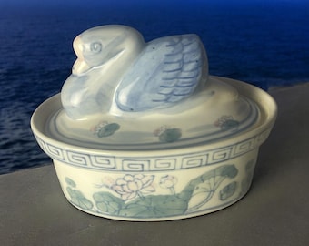 May include: A ceramic trinket box with a swan-shaped lid. The swan is white and blue, with detailed wing patterns. The box is white with blue and green floral designs and a decorative border. The background shows a blue ocean.