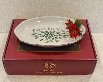 May include: White oval ceramic tray with gold trim and a green holly design. The tray says "Bless This Home" and has a red poinsettia flower on top. The tray is in its original box, which is red and says "Lenox Holiday Bless This Home Tray".