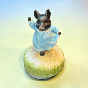 May include: A ceramic figurine of a dark gray piglet wearing a light blue dress with a white collar. The piglet is standing on a circular base painted to resemble a grassy mound, with one arm raised in a waving gesture and the dress flared out.