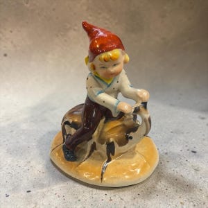 May include: A ceramic figurine of a small figure wearing a bright red pointed hat, a white long-sleeved shirt with small dark dots, and dark brown trousers. The figure is mounted on a brown and tan animal, all set on a round base.