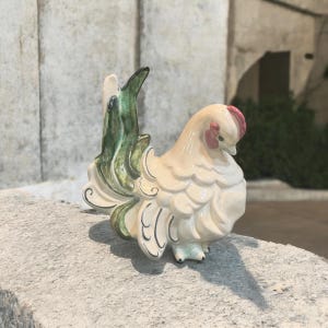 May include: A ceramic figurine of a rooster with a cream-colored body, a red comb, and a green and black tail. The rooster is perched on a stone surface. The figurine has a glossy finish and is likely a decorative item.