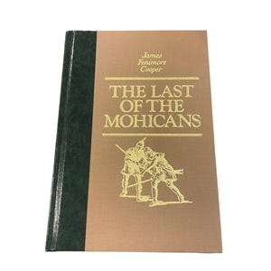 May include: A hardcover book titled "The Last of the Mohicans" by James Fenimore Cooper. The book has a tan cover with gold lettering and a green spine. The cover features an illustration of two figures in a fight.