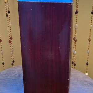 May include: A tall, rectangular box with a dark burgundy finish. The top edge is a contrasting blue. The box is positioned upright, suggesting it contains a product. The background includes decorative beaded strands.