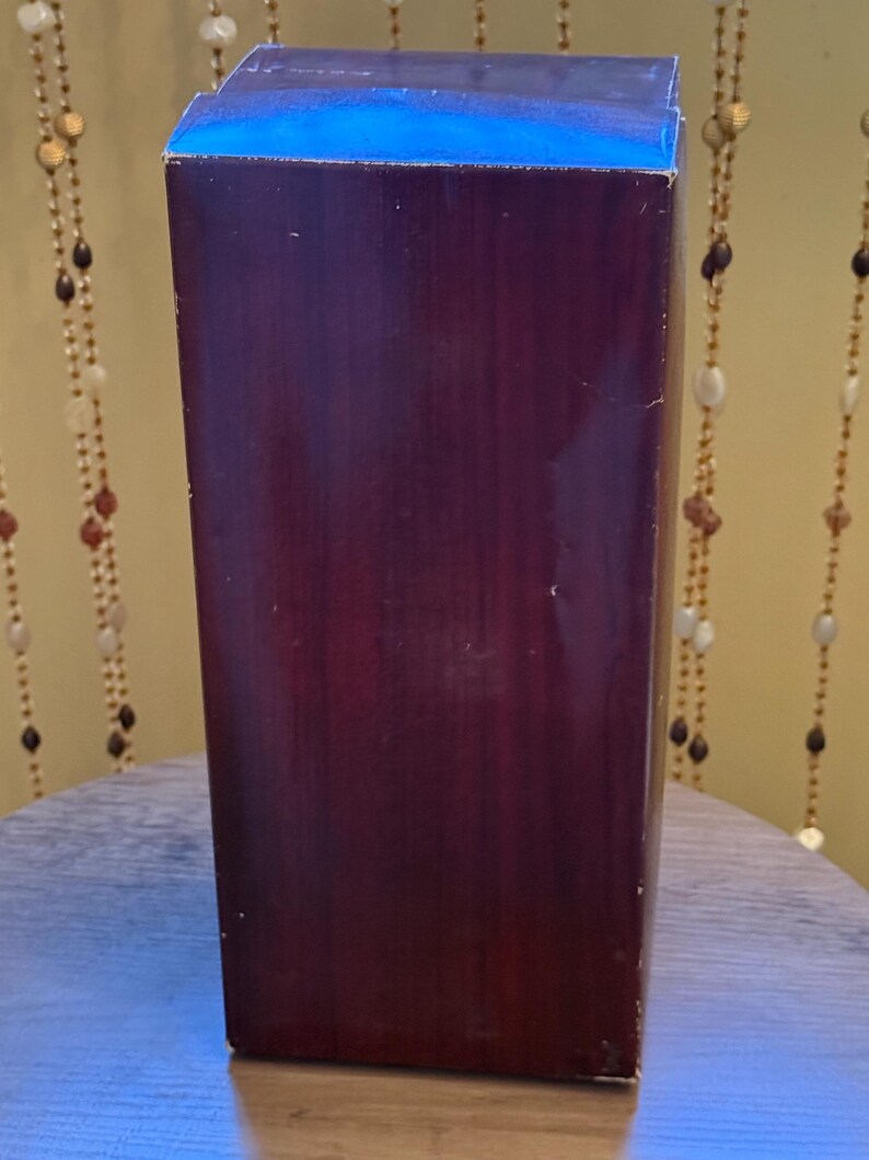 May include: A tall, rectangular box with a dark burgundy finish. The top edge is a contrasting blue. The box is positioned upright, suggesting it contains a product. The background includes decorative beaded strands.
