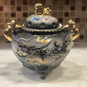 May include: A small, gray ceramic incense burner with gold accents and a dragon design. The burner has three legs and a lid with holes for incense sticks.