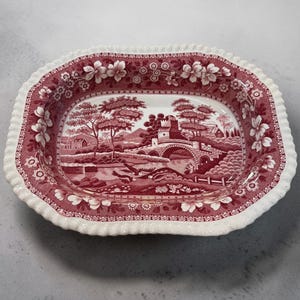 May include: A rectangular ceramic serving dish with a scalloped edge, featuring a deep red and white transferware pattern. The design depicts a landscape with a stone bridge, trees, and buildings, surrounded by a floral border on the rim.