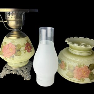 Puede incluir: Una l&aacute;mpara de aceite antigua con base de lat&oacute;n y pantalla de cristal pintada con motivos florales. La l&aacute;mpara incluye una chimenea de cristal transparente y una pantalla a juego separada. Las pantallas est&aacute;n pintadas con flores rosas y rojas y hojas verdes.