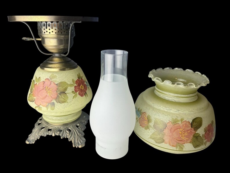 Puede incluir: Una l&aacute;mpara de aceite antigua con base de lat&oacute;n y pantalla de cristal pintada con motivos florales. La l&aacute;mpara incluye una chimenea de cristal transparente y una pantalla a juego separada. Las pantallas est&aacute;n pintadas con flores rosas y rojas y hojas verdes.