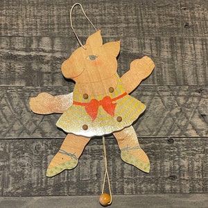 May include: A wooden pull-string puppet of a pig wearing a yellow and gold dress with a red bow. The puppet has a string attached to its feet and a wooden ball at the end of the string.