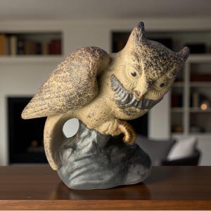 May include: A ceramic owl figurine with a detailed, textured design. The owl is perched on a dark gray rock, with its wings partially spread. The owl's feathers are a mix of tan and brown, with black accents. The figurine has a vintage or antique aesthetic.
