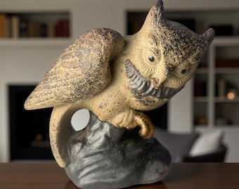 May include: A ceramic owl figurine with a detailed, textured design. The owl is perched on a dark gray rock, with its wings partially spread. The owl's feathers are a mix of tan and brown, with black accents. The figurine has a vintage or antique aesthetic.