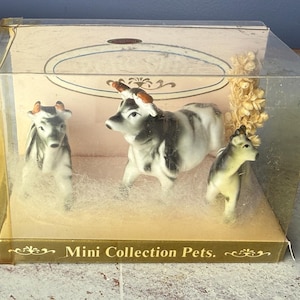 May include: A clear plastic box containing three miniature cow figurines. The cows are black and white with brown horns, set on a bed of white fluff. The box has the text "Mini Collection Pets."