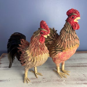 May include: Two handmade rooster figurines made of straw and fabric. The roosters are red, brown, and yellow with black feathers. They are standing on a white wooden surface.
