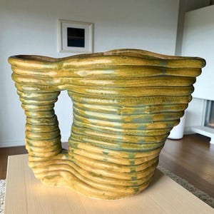 May include: A large, abstract ceramic sculpture with a unique, open design. The piece features horizontal, layered ridges in shades of yellow and green, creating a textured surface. The sculpture is placed on a light-colored wooden table.