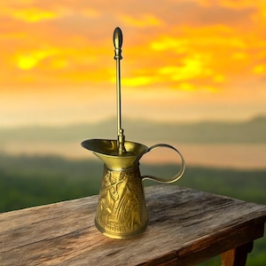 May include: A brass-colored decorative pitcher with a long, slender handle and a matching stirring rod. The pitcher features intricate embossed designs and a curved handle. The background shows a sunset over a landscape.