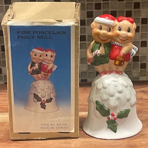 May include: A white porcelain bell with two pig figurines wearing Santa hats. The bell has a green holly sprig with red berries. The box reads "Fine Porcelain Piggy Bell" and "Item No. S22/6147 Made in Taiwan".
